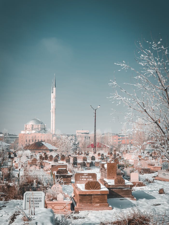 Winter scene of a cemetery covered in snow with a mosque in the distance under a clear blue sky.